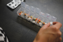 A hand placing a pill in a weekly pillbox with an assortment of daily medications